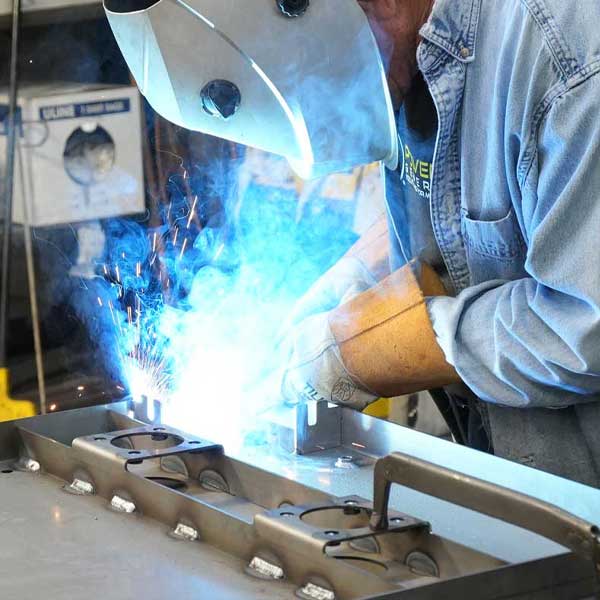 Welder using MIG or TIG welding techniques, sparks and blue flames visible, working on metal components for custom generator parts.